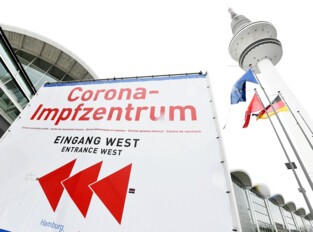 Hinweisschild mit Richtungsangabe zum Corona-Impfzentrum vor den Messehallen. Im Hintergrund der Fernsehturm.