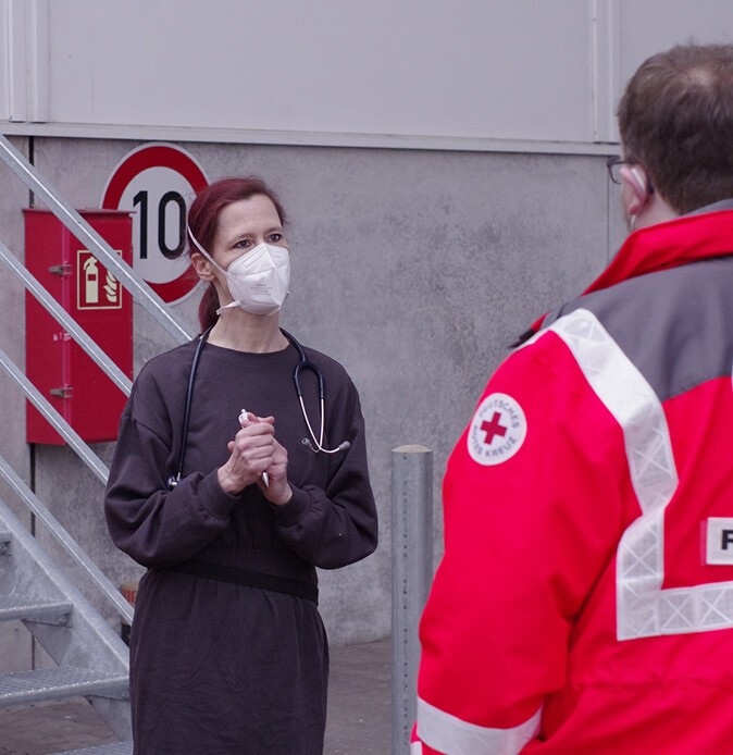 Eine Ärztin mit Mundschutz und Schutzkleidung spricht mit einem Mitarbeiter des deutschen roten Kreuzes