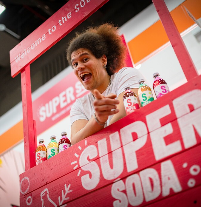 Ein lachender Mann hinter einem Messestand mit der Aufschrift Super Soda